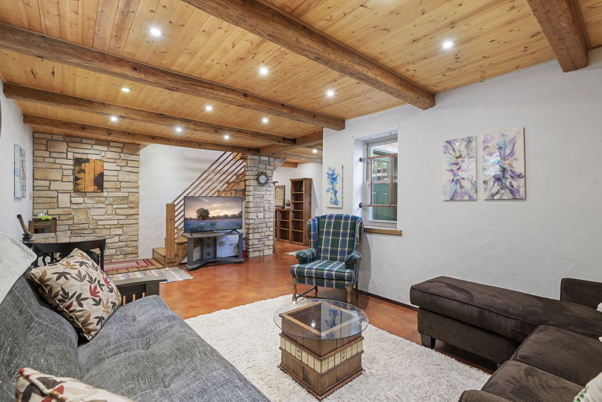 A spacious living area with a stone fireplace, wooden ceiling beams, and a plaid armchair.
