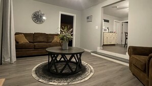 Un salon avec un ventilateur de plafond, une table ronde ornée d’une plante, un canapé et, au fond, un coin cuisine.