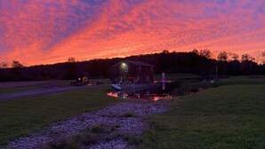 A rustic barn with a small shed and a picnic area, surrounded by a grassy field and a body of water. - Secluded Cabin w/ Private Pond/ Peaceful Getaway (Cameron Mills)