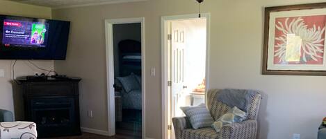 Una sala de estar con televisor, ventilador de techo y puerta que da a un dormitorio.