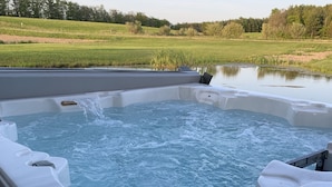 A hot tub with a wooden deck, surrounded by a grassy field and a body of water. - Secluded Cabin w/ Private Pond/ Peaceful Getaway (Cameron Mills)