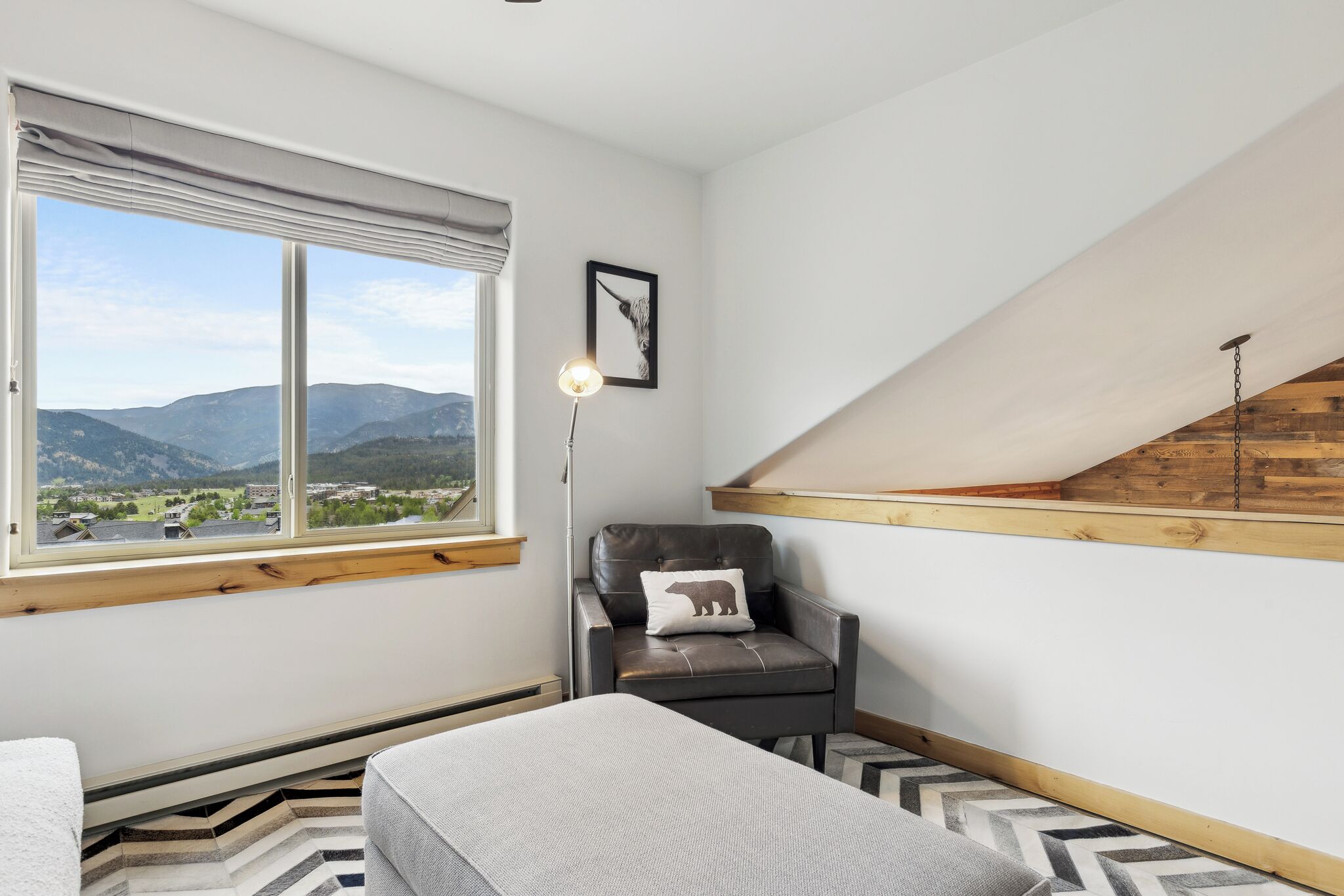 A room with a large window offering a mountain view, a leather chair with a bear pillow, a floor lamp, and a herringbone-patterned rug.
