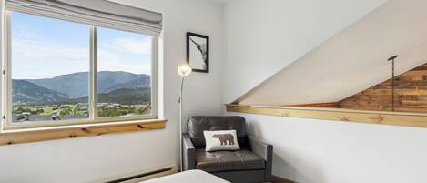 A room with a large window offering a mountain view, a leather chair with a bear pillow, a floor lamp, and a herringbone-patterned rug.