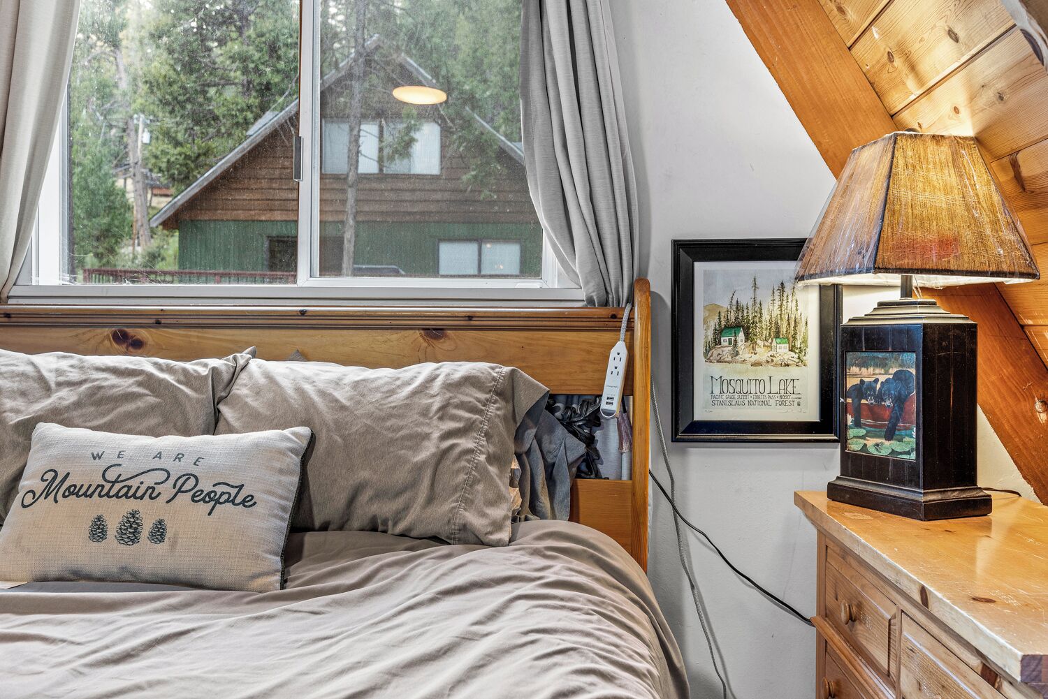 Un dormitorio con cama, lámpara, cuadro enmarco y ventana con vista a una casa.