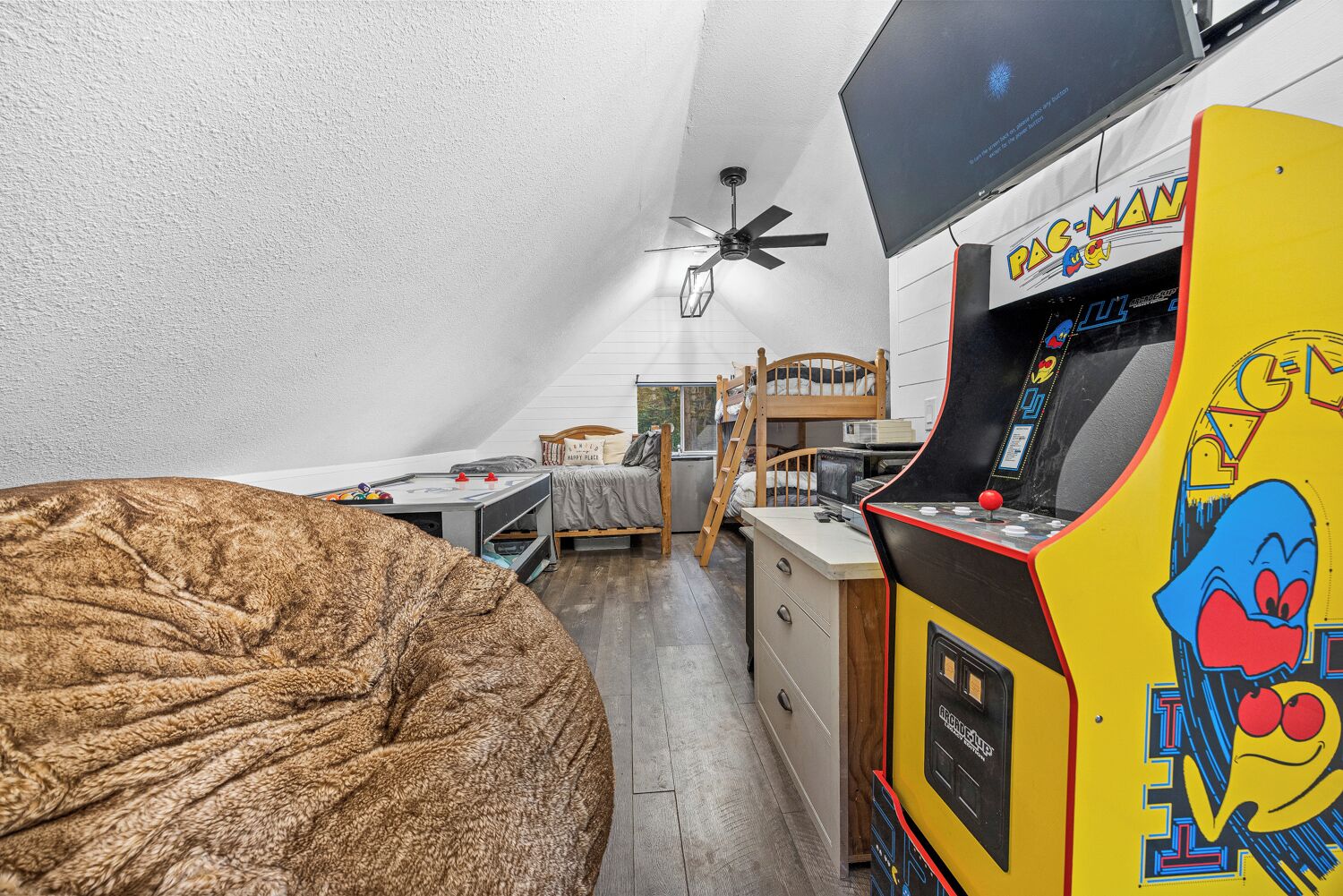 A compact room with a bunk bed, a desk with a computer, a large bean bag, and a Pac-Man arcade machine.