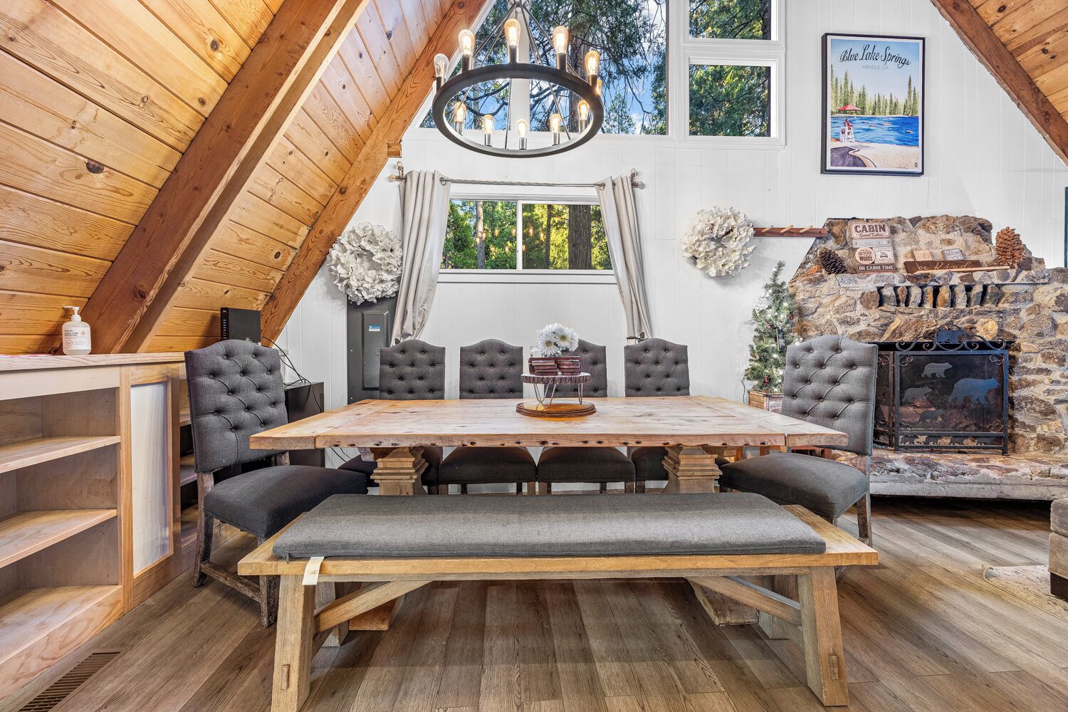 A cozy cabin interior with a wooden dining table, cushioned chairs, a fireplace, and a scenic view through the window.