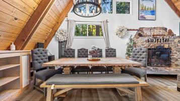 A cozy cabin interior with a wooden dining table, cushioned chairs, a fireplace, and a scenic view through the window.