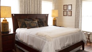 Una cama bien hecha con edredón blanco y varios almohadones, flanqueada por dos mesitas de noche con lámparas, en una habitación con paneles de madera y cuadros en la pared.