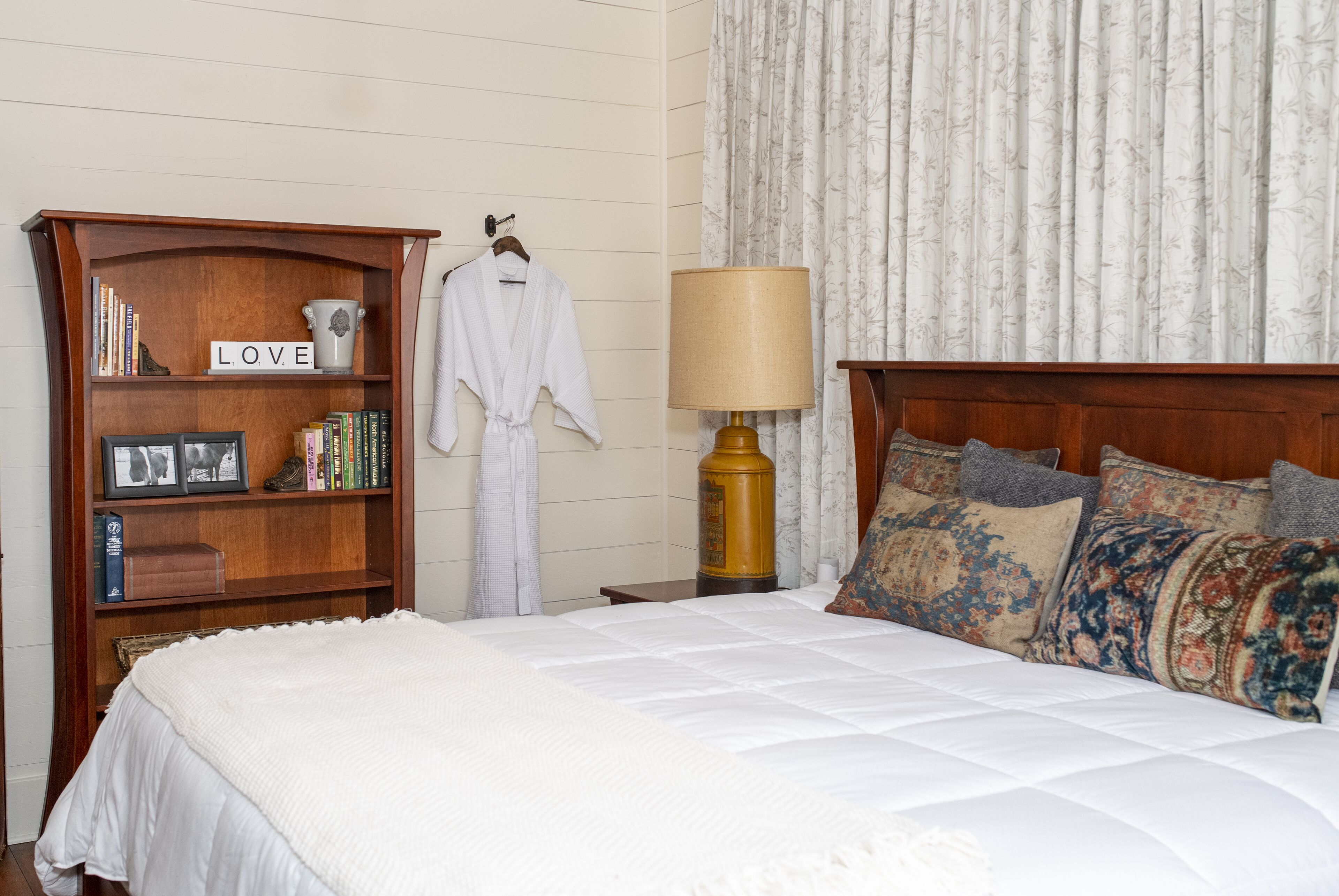 A neatly made bed with a white comforter and multiple decorative pillows, a wooden bookshelf with books and a 'LOVE' sign, a white bathrobe hanging on the wall, and a classic lamp on a bedside table.