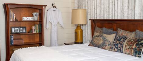 A neatly made bed with a white comforter and multiple decorative pillows, a wooden bookshelf with books and a 'LOVE' sign, a white bathrobe hanging on the wall, and a classic lamp on a bedside table.