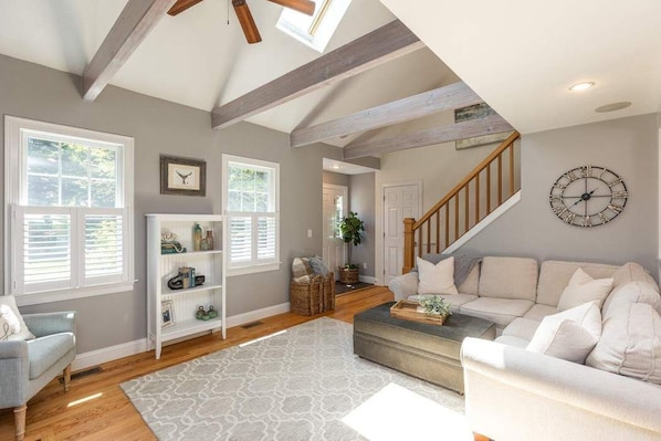 A spacious living room with a high ceiling, wooden beams, a staircase, and a large clock on the wall. - Vacation Getaway- Salt Water Pool, Firepit, Outdoor Bar/Kitchen. (Harwich)