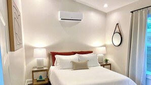 A neatly arranged bedroom with a bed, two bedside lamps, a wall-mounted air conditioner, and a window with curtains. - Modern guesthouse in downtown TR (Travelers Rest)