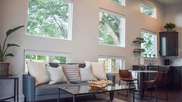A modern living room with a grey sofa, a glass coffee table, and a view of trees through large windows. - Modern guesthouse in downtown TR (Travelers Rest)