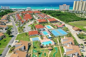 A resort with multiple buildings, a swimming pool, tennis courts, and a view of the ocean.