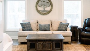 A living room with a beige sofa, patterned cushions, a wooden coffee table, and a wall-mounted tree artwork. - River View Loft on the Mermentau River with pool access (Mermentau)