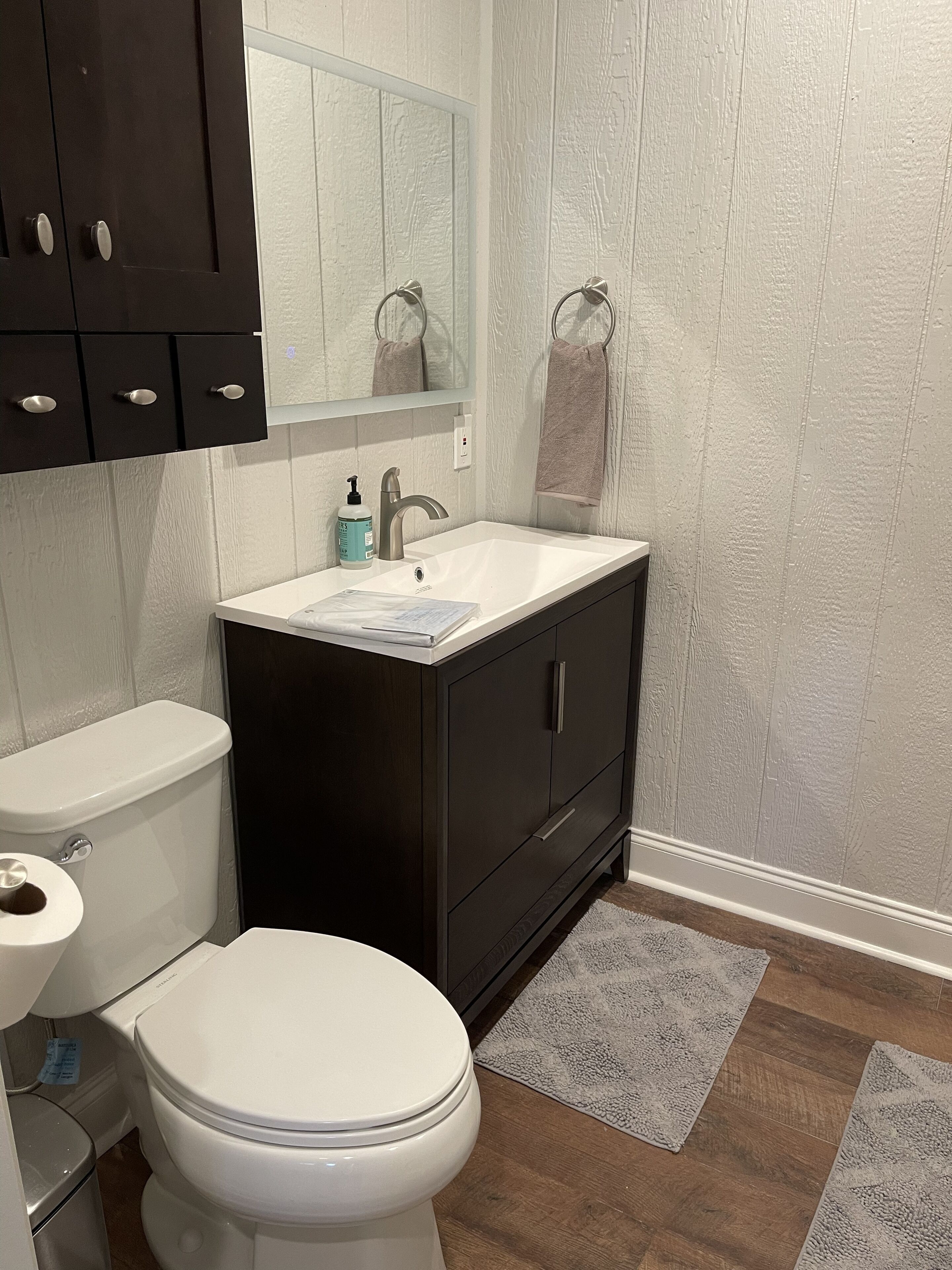 A bathroom with a white toilet, a dark wooden cabinet with a mirror above the sink, and a towel hanging on the wall.