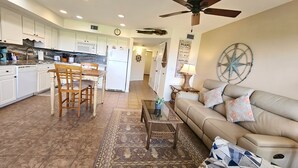 A spacious living area with a beige sofa, a coffee table, and a dining table with chairs. A ceiling fan is visible, and a wall-mounted clock is present. - Nature's Landing - A102 Budget Rates Downtown Unit (Cedar Key)