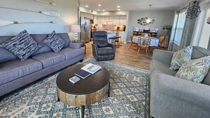A modern living room with a grey sofa, a round coffee table, and a dining area with a table and chairs. - Nature's Landing A104 Prettiest Condo in Town! (Cedar Key)