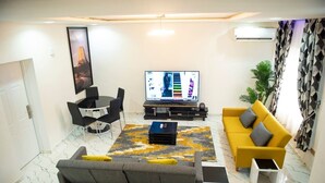 Uma sala de estar com sofá, um sofá amarelo, TV e uma mesa de jantar com cadeiras.