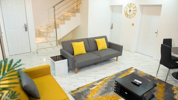 Uma sala de estar moderna com sofá cinzento, almofadas amarelas, mesa de centro preta e escada com corrimão de vidro.