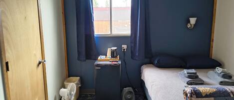 A small room with a bed, a desk with a fan, a window with curtains, and a door.