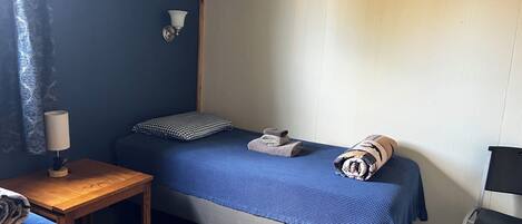 Una habitación pequeña con cama individual, colcha azul, mesita de noche con lámpara, una silla y cortina.