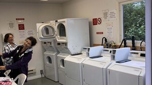Une laverie équipée de plusieurs machines à laver, une table recouverte d’une nappe rouge et des panneaux indiquant l’interdiction de fumer et les tarifs de lavage.