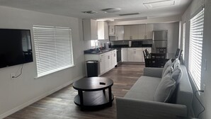 A modern living room with a sofa, a round coffee table, a TV, and a kitchen in the background. - Beach Ave Escape Unit B - Just steps from the Beach! (Redington Shores)