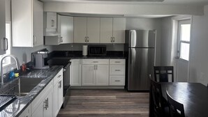 A modern kitchen with white cabinets, stainless steel appliances, a granite countertop, and a dining area with a dark wooden table and chairs. - Beach Ave Escape Unit B - Just steps from the Beach! (Redington Shores)