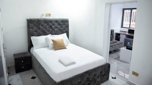 A neatly made bed with white linens and a single brown pillow, a dark tufted headboard, and a small bedside table. - Lovely One-bed Apartment Located in Abuja (Abuja)