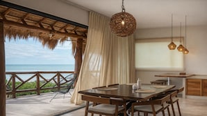 A dining area with a wooden table and chairs, a woven pendant light, and a view of the ocean through a thatched roof and curtains. - Casa Astral Hotel Boutique and Apartments (Isla Holbox)