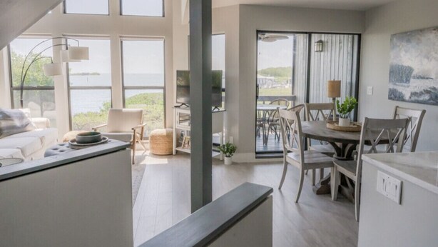 A spacious living area with a large window overlooking a body of water, a dining area with a table and chairs, and a modern kitchen with a counter. - CaptiVista! Your dream waterfront escape! Rustic luxury with unbelievable views! (North Captiva Island)