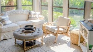 A spacious living room with a white sofa, a round ottoman, and a wooden chair. Large windows provide a view of greenery outside. - CaptiVista! Your dream waterfront escape! Rustic luxury with unbelievable views! (North Captiva Island)