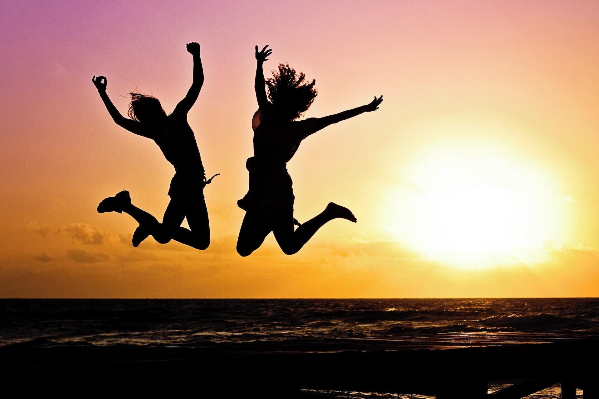Two silhouetted figures jumping against a sunset over the ocean.