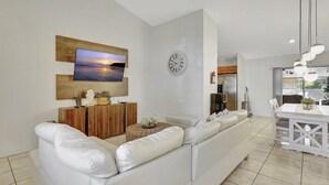 A modern living room with a white sofa, a flat-screen TV mounted on a wooden panel, a wall clock, and a dining area with a white table and chairs.
