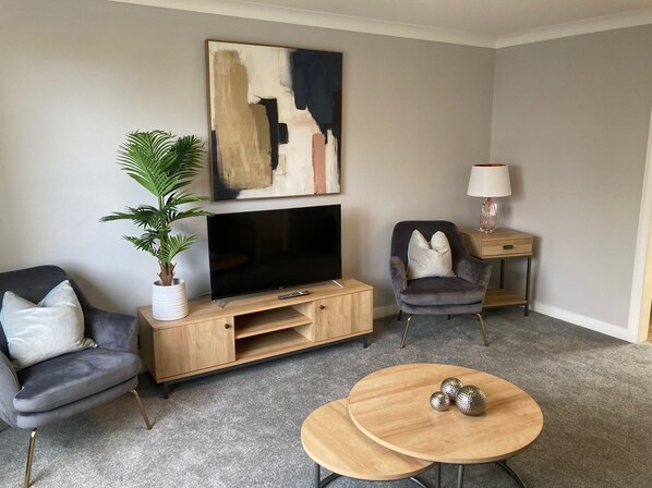 A modern living room with a flat-screen TV, two armchairs, a coffee table, a side table with a lamp, and a potted plant. - Leeward House - Donnini Apartments (Ayr)
