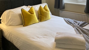 Una cama bien hecha con almohadas blancas y amarillas, un cabecero gris y una ventana con cortinas.