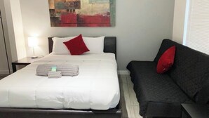 Ein ordentlich bezogenes Bett mit weißer Bettwäsche und einem roten Kopfkissen, ein schwarzes Sofa mit einem roten Kissen und ein Bild an der Wand.
