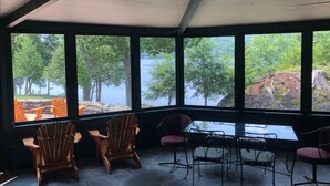 Un espace extérieur aménagé, abrité et doté de chaises en bois et d’une table, ainsi que de grandes fenêtres offrant une vue sur un lac et une forêt.