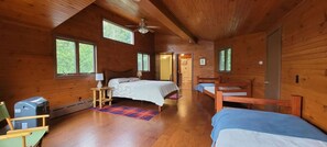 Un dormitorio de cabaña de madera con una cama, dos literas, un ventilador de techo y ventanas amplias.