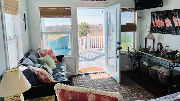 A living room with a sofa, a coffee table, a TV, and a balcony with a view of the landscape. - Home with beach view from  porch and it’s only a two minute walk to the beach. (Crystal Beach)