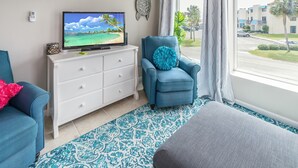 A living room with a TV, a blue armchair, a white dresser, and a view of a street with palm trees. - Comfy King beds, close to beach, pool + pet friendly  (Eastpoint)
