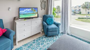 Un salon avec un téléviseur, un fauteuil bleu, une commode blanche et une vue sur une rue bordée de palmiers.