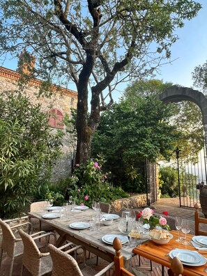 An outdoor dining area with a rustic wooden table set for a meal, surrounded by lush greenery and a stone building in the background.