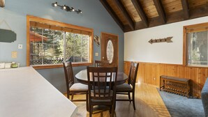 Un comedor con mesa y sillas de madera, ventanas amplias con vistas a árboles y techo de madera.