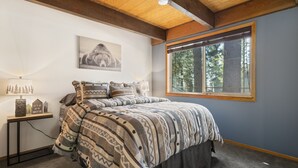 Un dormitorio con una cama, un ventanal con vistas a árboles, un cuadro en la pared y una mesita de noche con lámpara y objetos decorativos.