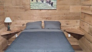 Una habitación de madera con una cama, dos mesitas de noche, una lámpara y un cuadro con ganado en la pared.