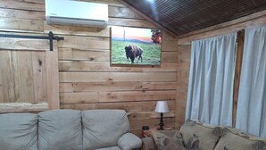 Un interior de madera con un sofá y una mesa, un cuadro de una vaca en la pared, y una ventana con cortinas.