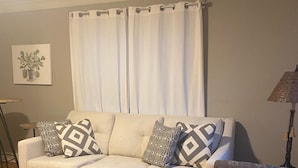 A living room with a beige sofa, patterned cushions, a dark patterned armchair, a wooden floor, a white curtain, and a small table with a plant. - Walking distance to Baum Stadium & Razorback Greenway access across the street! (Fayetteville)