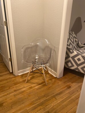 A clear plastic chair with wooden legs against a wall, wooden flooring, and a patterned sofa.